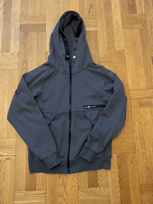 Grå hoodie med dragkedja från sail racing - Snygg grå hoodie från sail racing med hel dragkedja framtill och stor huva med tryckknappar och svart detaljband. Hoodien har ribbade muddar, två fickor och en liten logga på bröstet. Perfekt för en avslappnad och sportig stil.