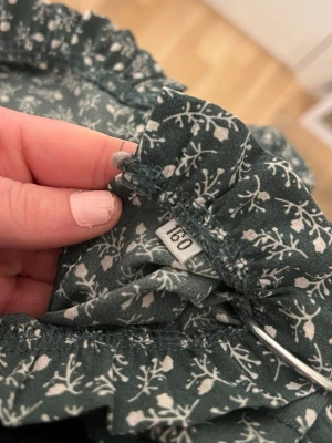 Grön blommig blus med volangkrage - Mörkgrön blus med vitt blommönster och volangdetaljer vid kragen. Långärmad modell i lätt och luftig kvalitet, perfekt för en trendig och avslappnad look. Passar dig som gillar romantiska detaljer och mjuka material.