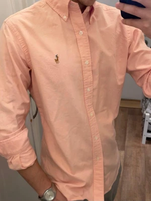 Rosa Skjorta från Ralph Lauren (Slim Fit) - Klassisk ljusrosa oxford skjorta från Polo Ralph Lauren med button-down krage och broderad logga på bröstet. Skjortan har långa ärmar, vita knappar och är tillverkad i mjuk bomull. Perfekt för en clean och stilren look. Storlek Small. Snabb Leverans 