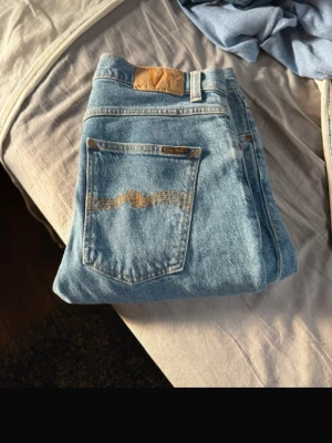 Blå jeans från Nudie Jeans - Säljer ett par klassiska blå jeans från Nudie Jeans med snygga kontrastsömmar och ikonisk våg på bakfickan. Jeansen har rak passform och är gjorda i slitstark denim. Perfekta för en avslappnad och trendig look.