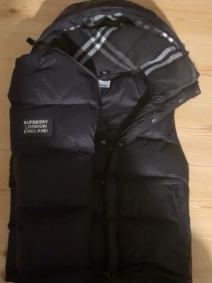 Svart Burberry Jacka - Snygg svart pufferjacka från Burberry med klassiskt rutigt innerfoder i huvan och patch med Burberry London England på bröstet. Jackan har stora vadderade partier, dragkedja och tryckknappar samt praktisk huva. Perfekt för kalla vinterdagar och västen är perfekt för den sköna vårtiden.