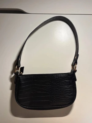 Svart axelväska i imiterad ormmönster - Snygg svart axelväska med imiterad ormskinns mönstrad yta och guldfärgade metalldetaljer. Väskan har en kort axelrem och en stilren, avlång form. Materialet är konstläder och gjord av polyester. Finns en innerficka med dragkedja. 
