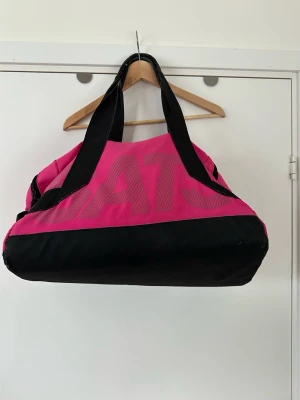 Rosa och svart träningsväska från SATS - Träningsväska från SATS i rosa och svart. Praktisk design med ett stort huvudfack och flera mindre fack med dragkedja på utsidan, samt ett skofack. Perfekt för gymmet eller som en mindre weekendväska. Fint skick!  Skickas mot porto, kan även hämtas i Stockholm. 