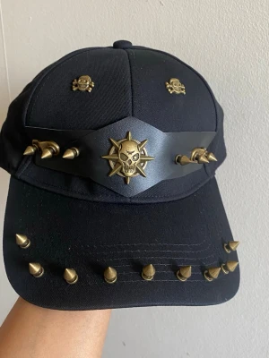 Svart keps med nitar och dödskallar - Säljer en svart keps med coola guldfärgade nitar och dödskalle-detaljer. Kepsen har en unik design med metallspikar på skärmen och ett band med dödskalleemblem framtill. Perfekt för dig som gillar alternativ stil och vill sticka ut.