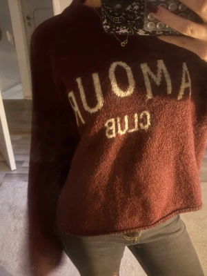 Vinröd stickad tröja med text - Mysig vinröd stickad tröja med texten 'AMOUR CLUB' i vitt framtill. Tröjan har en avslappnad passform, långa ärmar och mjukt material som passar perfekt till höst och vinter. Enkel att styla med jeans eller kjol för en chill look.