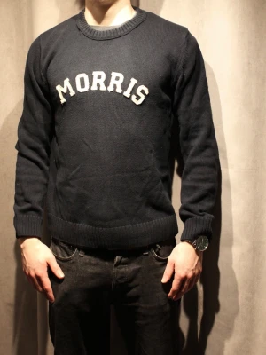 Marinblå stickad tröja Morris - Snygg marinblå stickad tröja från Morris med vit broderad logga på bröstet. Tröjan har rund halsringning, ribbade muddar vid ärmslut och nederkant samt en klassisk, rak passform. Perfekt för en stilren och avslappnad look.