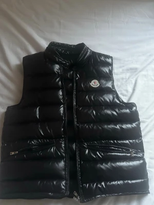 Svart dunväst från Moncler - Snygg svart dunväst från Moncler med glansig finish och diskret logga på bröstet. Västen har hög krage, två dragkedjeförsedda fickor och stängs med dragkedja framtill. Tillverkad i dun och certifierad enligt DIST-systemet. OBS, fixat hål på baksidan av västen som knappast syns