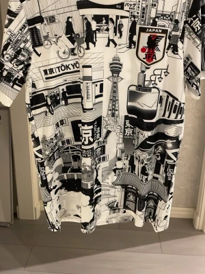 Japan T-shirt med Tokyo-motiv - Cool svartvit t-shirt från Japan FA, adidas med detaljerade tryck av Tokyo och japanska stadsmotiv. T-shirten har korta ärmar, rund hals och är tillverkad i mjuk bomull. Perfekt för dig som gillar streetstyle och unika prints.