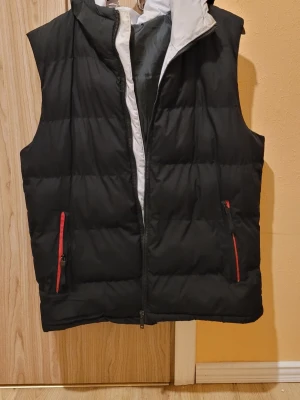 Svart dunväst med huva och röda detaljer - Snygg svart dunväst med avtagbar huva och röda dragkedjor på fickorna. Västen har quiltad design och vit insida vid kragen. Perfekt för lager-på-lager och håller dig varm under kyliga dagar.