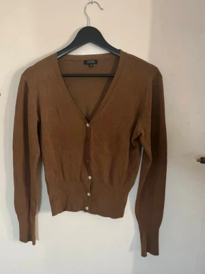 Brun kofta med knappar  - Snygg brun kofta med v-ringning och vita knappar framtill. Modellen är figurnära med långa ärmar och ribbade muddar. Perfekt att matcha med jeans eller kjol för en trendig look. Skönt tyg och sitter bra på. Normal i storleken och har en bra passform. 