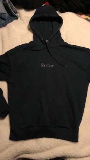 Mörkblå hoodie från Solid heritage  - Snygg mörkblå hoodie från Solid med texten 'Évoluer' tryckt på bröstet. Klassisk modell med huva och dragsko, samt ribbade muddar vid ärmar och nederkant. Perfekt för en avslappnad och trendig stil.