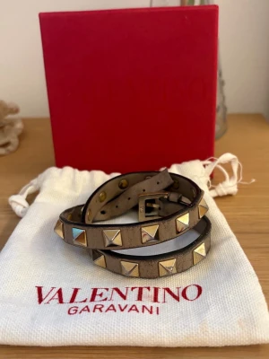 Valentino Garavani nitarmläderarmband - Snyggt brunt ish läderarmband från Valentino. Armbandet kan lindas två varv runt hand leden. Haft i cirka 1 år. Sönder längst ut men spelar egentligen ingen roll när man spänner ihop armbandet förutom att de kan vara lite svårt. 