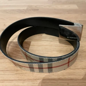 Burberry bälte med klassiskt rutmönster - Snyggt bälte från Burberry med det ikoniska beige, röda och svarta rutmönstret. Bältet har en silverfärgad metallspänne med Burberry-logga och är gjort i läder. Perfekt accessoar för att lyfta vilken outfit som helst.