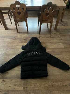 Svart pufferjacka från Tullsta - Svart pufferjacka med huva från Trapstar Jackan har dragkedja framtill, två fickor och broderad logga på bröstet. På ryggen står Trapstar i stora bokstäver. Perfekt för kalla dagar och riktigt skön passform.