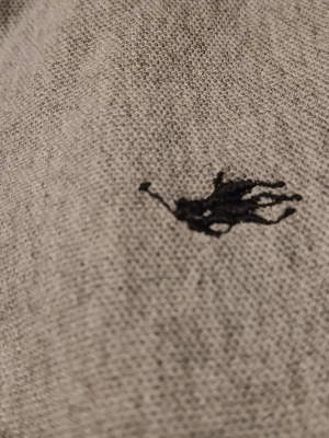 Grå polotopp från Polo Ralph Lauren - Klassisk grå polotopp från Polo Ralph Lauren med korta ärmar och krage. Toppens material är mjuk bomull och den har knappar framtill samt den ikoniska logon broderad på bröstet. Perfekt för en avslappnad och stilren look.