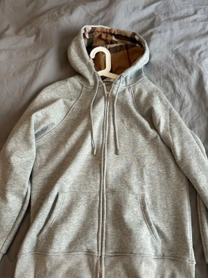 Grå zip hoodie från Burberry - Använd 3 gånger! Snygg grå zip hoodie från Burberry med klassiskt rutigt foder i huvan och dragsko. Broderad logga på bröstet och två fickor framtill. Tillverkad i mjuk bomullsblandning som känns riktigt skön att ha på sig. Perfekt för en avslappnad och stilren look.