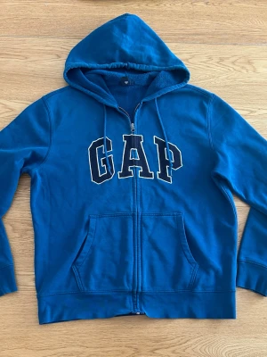 Blå zip hoodie från GAP - Blå hoodie från GAP med dragkedja och stor logga i svart och vitt på bröstet. Mjuk fleece på insidan, två fickor framtill och justerbar huva med snören. Perfekt för en avslappnad och cool stil.