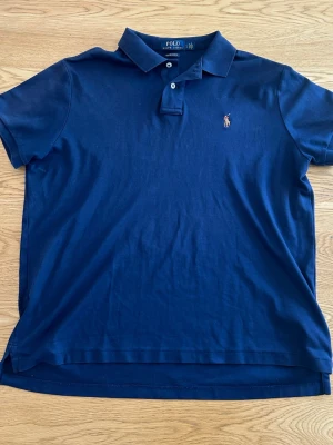 Marinblå piké från Polo Ralph Lauren - Säljer en marinblå pikétröja från Polo Ralph Lauren med klassisk krage, två knappar och den ikoniska broderade loggan på bröstet. Modellen är custom slim fit och tillverkad i mjuk bomull. Perfekt för en clean och stilren look.
