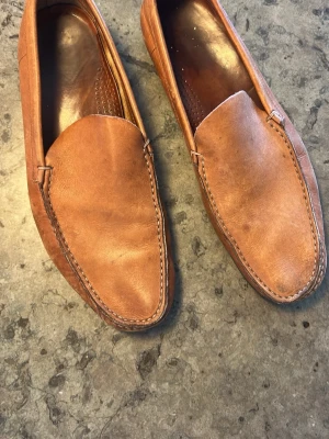 Bruna loafers i läder från Church’s. Nypris 7000 - Snygga bruna loafers i äkta läder med klassisk design och rundad tå. Skorna har dekorativa sömmar runt ovandelen och en stilren look som passar till många outfits. Perfekta för dig som gillar en tidlös och avslappnad stil. 
