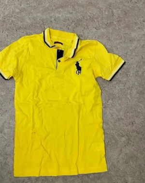 Gul piké t-shirt från Ralph Lauren - Snygg gul piké t-shirt från Ralph Lauren med klassisk krage och svart logga på bröstet. T-shirten har korta ärmar och svarta detaljer vid krage och ärmslut. Perfekt för en avslappnad men stilren look.
