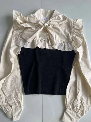 Krämvit blus med volanger och rosett - Trendig blus med krämvita puffärmar, stor rosett vid halsen och volangdetaljer över axlar och bröst. Kroppen är svart och ribbad vilket ger en snygg kontrast. Långa ärmar med knappar vid mudden. Perfekt för dig som gillar statement-plagg. 💕