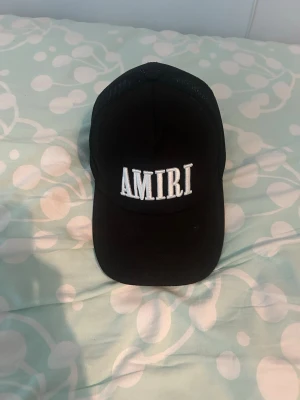 Svart keps från Amiri med mesh - Svart keps från Amiri med vit broderad logga framtill. Klassisk trucker-modell med böjd skärm och mesh på baksidan för extra ventilation. Justerbar passform baktill. Tillverkad i bomull och polyester.