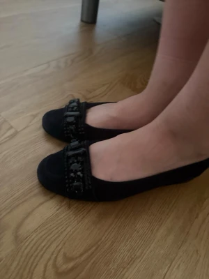 Svarta ballerinaskor med dekor - Säljer ett par svarta ballerinaskor med dekorativa detaljer framtill. Skorna har rundad tå och en snygg lackad dekoration på ovandelen. Perfekta för dig som vill ha en klassisk men ändå trendig look.