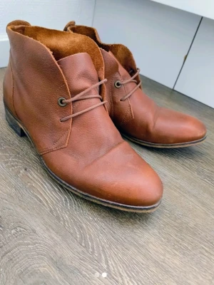 Bruna läderstövletter med snörning - Snygga bruna stövletter i äkta läder med klassisk snörning och låg klack. Skorna har en stilren design med rundad tå och råa kanter upptill. Perfekta för dig som gillar en tidlös och trendig look.