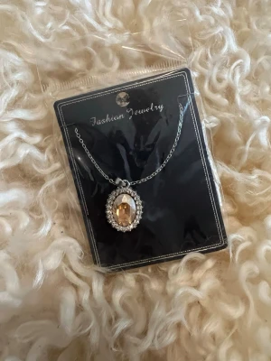 Silverfärgat halsband med glittrig sten - Snyggt halsband med tunn silverfärgad kedja och ett ovalt hänge med en stor, glittrande guldfärgad sten i mitten. Hänget är dekorerat med små klara stenar runt om som ger extra bling. Perfekt accessoar för att lyfta din outfit.