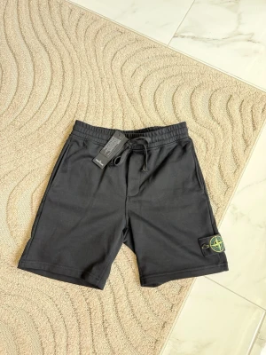 Svarta shorts från Stone Island - Svarta shorts från Stone Island med klassisk logopatch på benet. Modellen har elastisk midja med snörning och är tillverkad i mjuk bomull. Perfekta för en avslappnad och stilren look.