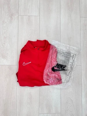Röd Nike Dri-FIT träningströja - Snygg röd träningströja från Nike med halv dragkedja och vit broderad logga på bröstet. Tillverkad i Dri-FIT-material som transporterar bort fukt och håller dig torr under träningen. Perfekt för sport eller gympass.