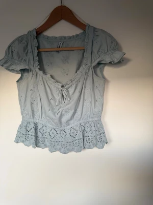 Ljusblå broderad blus med volang - Ljusblå blus i bomull med broderade blommor, korta puffärmar och volangkant nedtill. Toppen har v-ringning med knyt framtill och romantiska detaljer som spets och broderi.