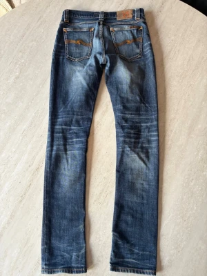 Blå smala jeans från Nudie Jeans - Säljer ett par blå jeans från Nudie Jeans med klassisk femficksmodell och slitna detaljer. Jeansen har kontrastsömmar och broderade fickor bak. Tillverkade i denim med snygg tvätt och lätt stretch för skön passform. Pris ej hugget i sten 
