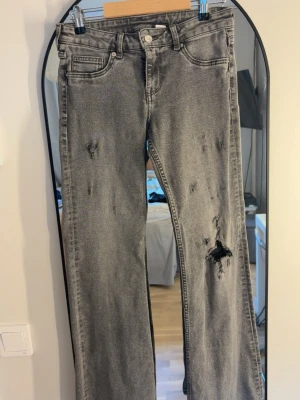 Grå slitna utsvängda jeans - Säljer ett par grå jeans med slitningar på både fram- och baksida. Jeansen har fem fickor, knappgylf och råa kanter vid bensluten för en trendig look. Perfekta för dig som gillar en avslappnad och edgy stil.