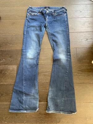 Blå bootcut jeans från True Religion - Säljer ett par blå bootcut jeans från True Religion med klassiska kontrastsömmar och snygga fickdetaljer bak. Jeansen har låg midja, slitningar framtill och breda ben. Perfekta för dig som gillar en avslappnad men trendig look. De är lite slitna längds ner men de ser man inte för de är på baksidan.