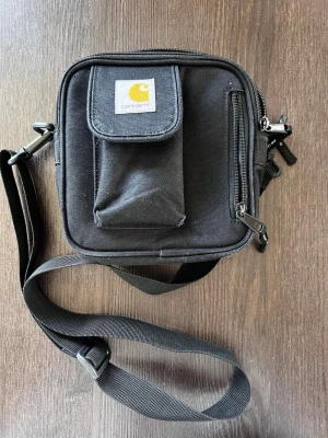 Svart axelväska från Carhartt - Snygg svart axelväska från Carhartt i slitstark canvas. Väskan har justerbar axelrem, en ficka med lock framtill och en dragkedjeförsedd ficka på framsidan. Perfekt för att bära det viktigaste och har en clean, fyrkantig form.