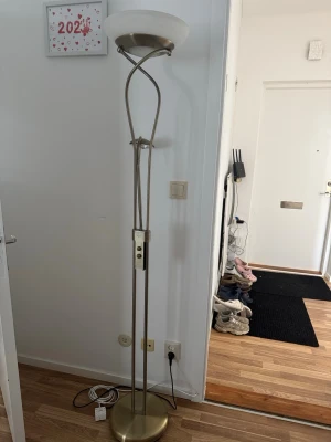 Golvlampa i mässing och glas - Snygg golvlampa med rund fot i mässingfärgad metall och vit glasskärm. Lampan har en modern design med böjda metallstavar och inbyggd dimmer på stativet. Perfekt för att skapa mysig belysning i vardagsrummet.
