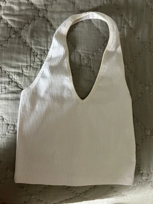 Vit ribbad halterneck linnetopp - Säljer en vit ribbad linnetopp med halterneck och djup v-ringning. Toppen är ärmlös och croppad, perfekt för varma dagar eller som layering piece. Materialet är mjukt och stretchigt för en bekväm passform.