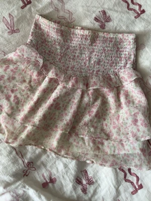 Blommig volangkjol i pastellrosa - Supersöt kortkjol med blommigt mönster i ljusrosa och vitt. Kjolen har bred smock i midjan och dubbla volanger för extra flowy känsla. Perfekt för vår och sommar! Materialet känns lätt och luftigt.