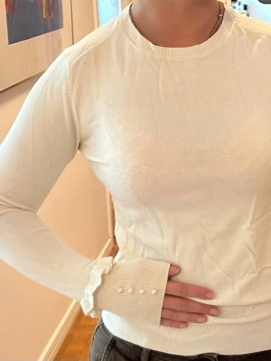 Vit stickad tröja med pärldetaljer - Snygg vit stickad tröja med långa ärmar och ribbade muddar. Ärmsluten har volangkant och dekorativa pärlor som ger en extra touch. Tröjan har rund halsringning och är figurnära i modellen. Perfekt för dig som gillar stilrena detaljer. Storlek M men passar även S
