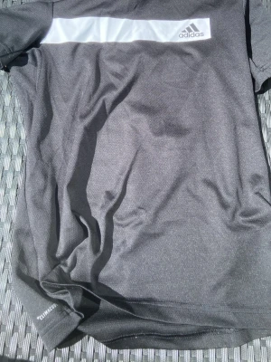 Svart tränings-tshirt från Adidas - Säljer en svart tränings-tshirt från Adidas med vit panel och logga på bröstet. Tillverkad i lätt och snabbtorkande funktionsmaterial, perfekt för träning eller sport. Kort ärm och klassisk passform.