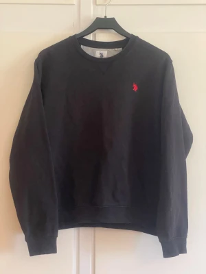 Svart sweatshirt från Polo Assn - Cool svart sweatshirt från Polo Assn med klassisk rund hals och röd broderad logga på bröstet. Tröjan har långa ärmar och ribbade muddar vid ärmslut och nederkant. Perfekt för en avslappnad och stilren look.