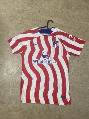 Atletico Madrid matchtröja röd/vit - Säljer en Atletico Madrid matchtröja från Nike med röda och vita vågiga ränder, klubbmärke på bröstet och sponsorloggor på fram- och baksidan. Tröjan har korta ärmar och är tillverkad i ett lätt, ventilerande material som passar perfekt till fotbollsträning.