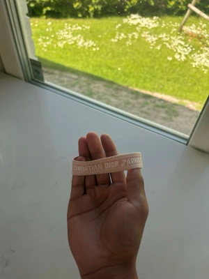 Christian Dior J'Adior armband.Stort Lager!!! - Snyggt armband från Christian Dior med texten 'J'Adior'. Finns i flera färger som ljusrosa, blå och svart. Armbandet är gjort i tyg med broderad text och har justerbara tofsar för perfekt passform. Perfekt accessoar för att lyfta din outfit.