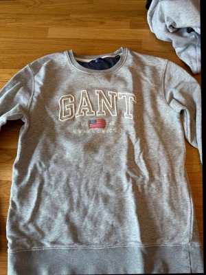 Grå sweatshirt från GANT - Snygg grå sweatshirt från GANT med broderad logga och amerikansk flagga på bröstet. Rund halsringning och mjukt material, perfekt för en avslappnad stil. Klassisk collegekänsla och tidlös design.