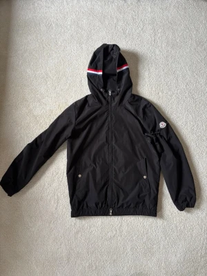 Svart Moncler vindjacka med huva - Snygg svart vindjacka från Moncler med huva och dragkedja framtill. Jackan har Moncler-logga på ärmen och rödvitblå rand på huvan. Två fickor med tryckknappar och elastisk nederkant. Perfekt för dig som vill ha en stilren och sportig look.