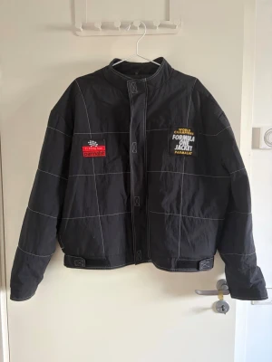 Svart Formula 1 bomberjacka - Svart bomberjacka med vita rutmönster och broderade Formula 1-detaljer från Parmalat. Jackan har hög krage, kardborreband vid ärmslut och midja samt dragkedja framtill. Perfekt för dig som gillar racing och streetwear.