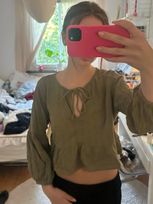 Olivgrön blus med knytning - Trendig olivgrön blus i luftig bomull med volangkant nedtill och knytning vid halsen. Blusen har långa, puffiga ärmar och en avslappnad passform som ger en bohemisk vibe. Perfekt att matcha med höga jeans eller kjol.