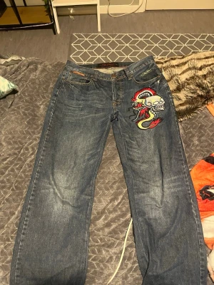 Ed Hardy jeans - Säljer ett par blå jeans från Ted Red med ett stort färgglatt dödskalle- och ormtryck på ena benet. Jeansen har raka ben, klassisk femficksmodell och coola detaljer som kontrastsömmar och metallknapp med krona. Perfekta för dig som vill sticka ut!