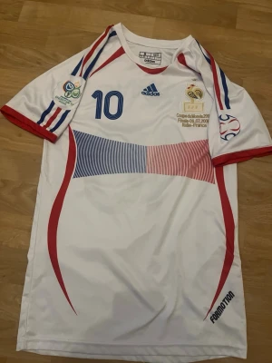 Frankrike fotbollströja Adidas S - Säljer en vit Frankrike fotbollströja från Adidas med nummer 10, blå och röda detaljer samt FFF-emblem. Tröjan har tryck från VM-finalen 2006 mot Italien och klassiska Adidas-ränder på ärmarna. Tillverkad i lätt och ventilerande material, perfekt för fotboll eller samling. Kan diskutera om pris 